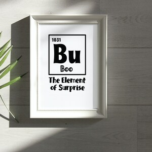 K&ouml;nnte beinhalten: Ein wei&szlig;er gerahmter Druck mit schwarzem Text, der "1031 Bu Boo The Element of Surprise" lautet. Der Text ist so gestaltet, dass er wie ein Element des Periodensystems aussieht.