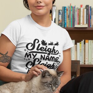 Puede incluir: Camiseta blanca con un gr&aacute;fico negro que dice "Sleigh My Name, Sleigh" con una silueta de Santa Claus y renos.