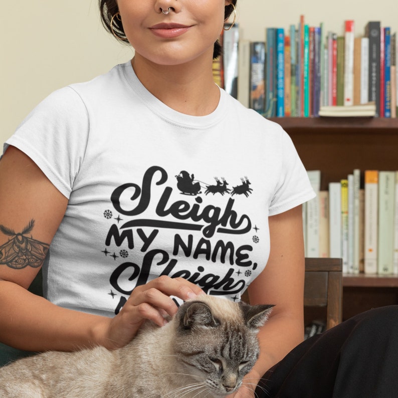 Puede incluir: Camiseta blanca con un gr&aacute;fico negro que dice "Sleigh My Name, Sleigh" con una silueta de Santa Claus y renos.