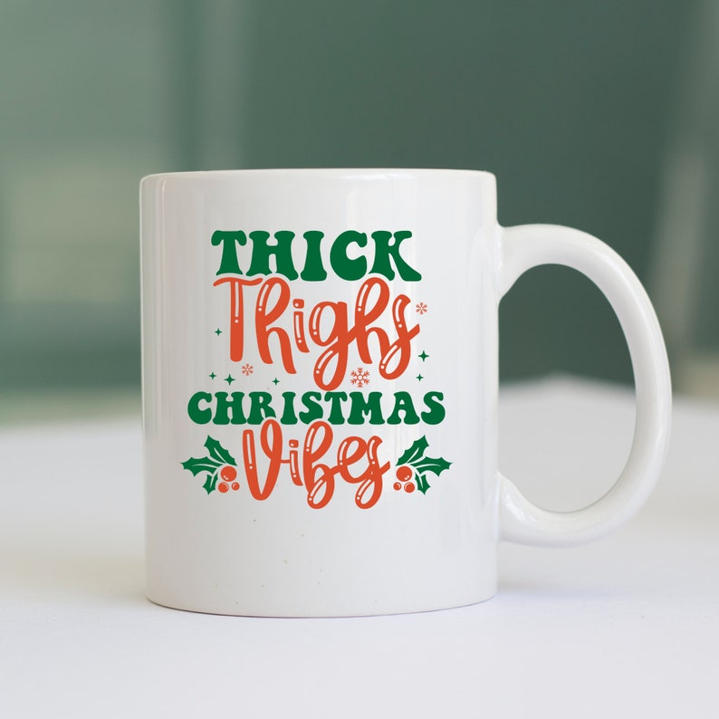 Puede incluir: Taza de cer&aacute;mica blanca con un dise&ntilde;o verde y rojo. El texto en la taza dice "Thick Thighs Christmas Vibes".