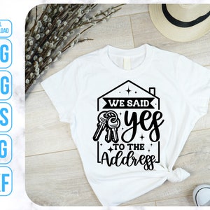 Op de afbeelding: Wit T-shirt met een zwart grafisch ontwerp van een huis met sleutels en de tekst "We said yes to the address".
