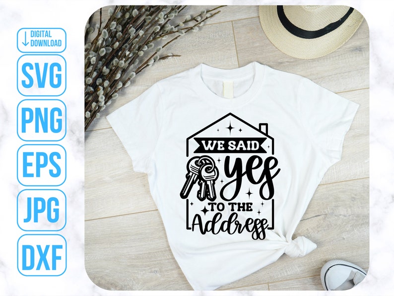 Op de afbeelding: Wit T-shirt met een zwart grafisch ontwerp van een huis met sleutels en de tekst "We said yes to the address".