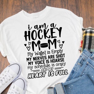 以下が含まれることがあります： 黒い文字で「i am a HOCKEY MOM」と書かれた白いTシャツ。 「M」の間にホッケーのパックがあります。 テキストは「My Wallet is Empty MY NERVES ARE SHOT MY VOICE IS HOARSE my schedule is crazy AND MY HEART IS FULL」と続きます