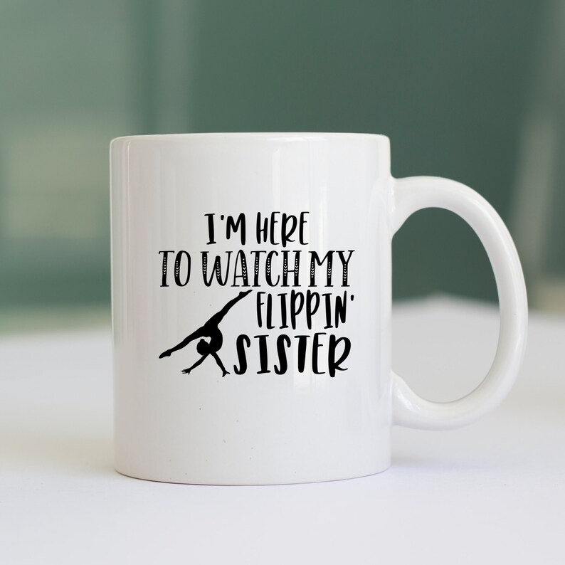 May include: White ceramic coffee mug with a black silhouette of a gymnast doing a back handspring. The text on the mug reads "I'm here to watch my flippin' sister."