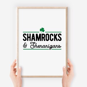 Op de afbeelding: Een witte ingelijste print met zwarte tekst die "Shamrocks & Shenanigans" luidt met een groene klaver boven de tekst.