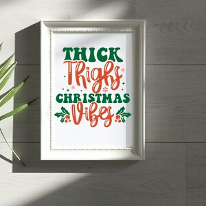 Puede incluir: Impresi&oacute;n enmarcada en blanco con el texto "Thick Thighs Christmas Vibes" en letras verdes y naranjas con acebo y copos de nieve.