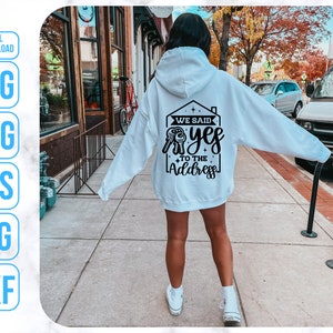 Op de afbeelding: Een witte hoodie met een zwarte afbeelding op de rug die "We said yes to the address" zegt. De afbeelding toont een huis met een sleutelgat en een sleutel.