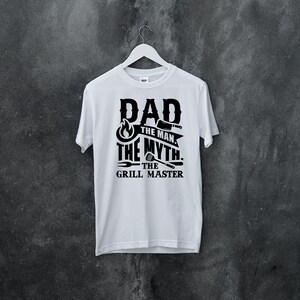 Puede incluir: Camiseta blanca con texto negro que dice "Dad The Man The Myth The Grill Master" con un gr&aacute;fico de llama y herramientas de parrilla.
