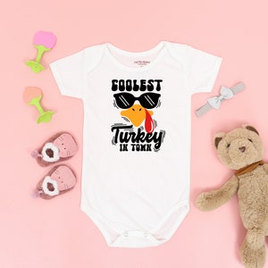 May include: White baby onesie with a cartoon turkey wearing sunglasses and the text "Coolest Turkey in Town".