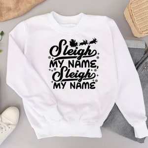 Puede incluir: Sudadera blanca con texto negro que dice "Sleigh my name, Sleigh my name" con una silueta de Pap&aacute; Noel en un trineo tirado por renos sobre el texto.
