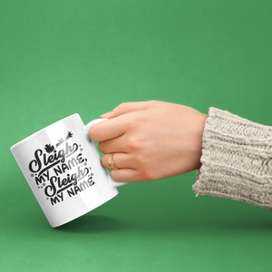 Puede incluir: Taza de cer&aacute;mica blanca con texto negro que dice "Sleigh My Name, Sleigh My Name". La taza est&aacute; siendo sostenida por una mano que lleva un su&eacute;ter color crema.