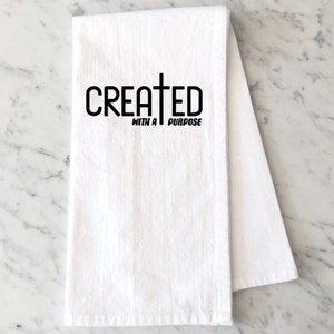 以下が含まれることがあります： 黒い文字で「CREATED WITH A PURPOSE」と十字架のシンボルが書かれた白いキッチンタオルです。