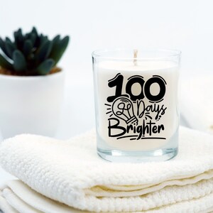 May include: A clear glass candle jar with a white candle inside. The candle has a black design that says "100 Days Brighter" with a lightbulb graphic.