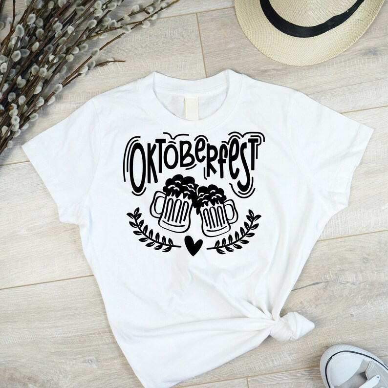 May include: White t-shirt with a black graphic design that says "Oktoberfest" with two beer mugs clinking and a heart in between.