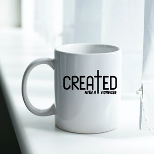 以下が含まれることがあります： 黒い文字で「CREATED WITH A PURPOSE」と黒十字が書かれた白いセラミックマグカップ