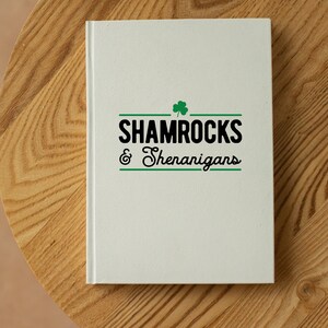 Op de afbeelding: Een wit notitieboek met een groene klaver en de tekst "Shamrocks & Shenanigans" in zwart.