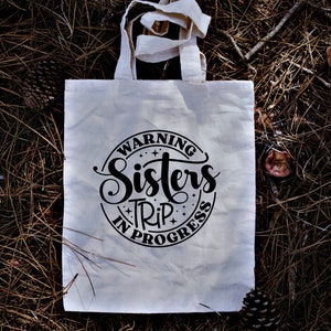 Puede incluir: Bolsa de tela blanca con texto negro que dice "Warning Sisters Trip in Progress" en un c&iacute;rculo con estrellas.