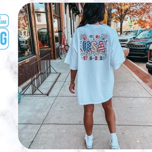 May include: A white t-shirt featuring a colourful graphic on the back with the text "USA freedom 1776-2023". The design incorporates stars, stripes, and fireworks. The shirt is worn by a person walking on a pavement.
