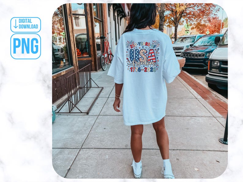 May include: A white t-shirt featuring a colourful graphic on the back with the text "USA freedom 1776-2023". The design incorporates stars, stripes, and fireworks. The shirt is worn by a person walking on a pavement.