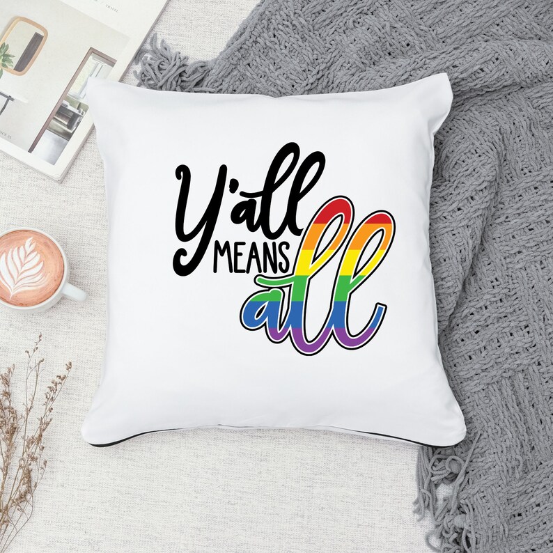 May include: White throw pillow with black text that reads "Y'all means all" with a rainbow arc behind the word "all".