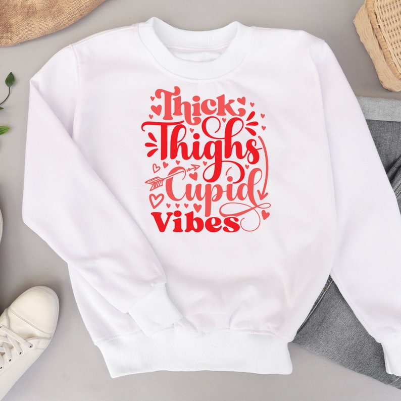 May include: White sweatshirt with red text that reads "Thick Thighs Cupid Vibes" surrounded by small red hearts.