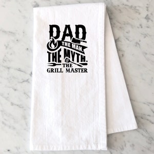 Puede incluir: Toalla de cocina blanca con texto negro que dice "Dad The Man The Myth The Grill Master" con un gr&aacute;fico de llama, cuchillo, esp&aacute;tula y tenedor.