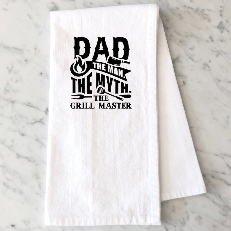 Puede incluir: Toalla de cocina blanca con texto negro que dice "Dad The Man The Myth The Grill Master" con un gr&aacute;fico de llama, cuchillo, esp&aacute;tula y tenedor.