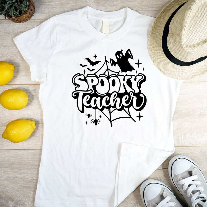 Pu&ograve; includere: Maglietta bianca con un design grafico nero di un fantasma, una ragnatela e pipistrelli. Il testo "Spooky Teacher" &egrave; scritto in un font stilizzato.