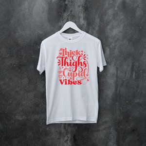 May include: White t-shirt with a red graphic that says "Thick Thighs Cupid Vibes" with hearts and arrows around the text.