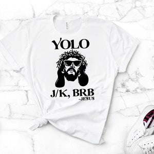 May include: White t-shirt with a black graphic of Jesus wearing sunglasses and a crown of thorns. The text "YOLO J/K, BRB -JESUS" is printed below the graphic.
