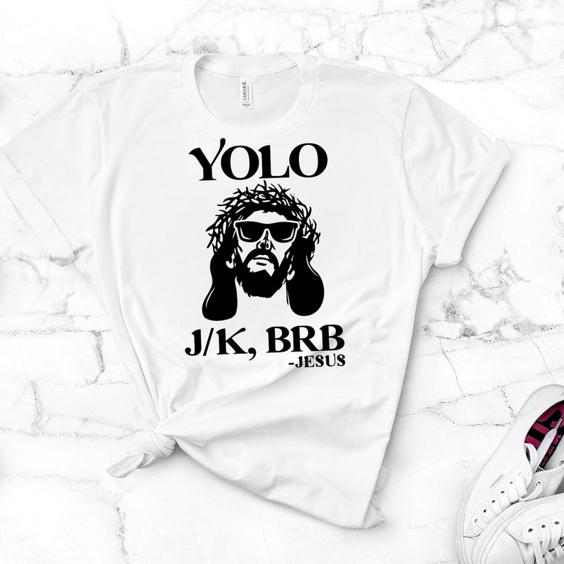May include: White t-shirt with a black graphic of Jesus wearing sunglasses and a crown of thorns. The text "YOLO J/K, BRB -JESUS" is printed below the graphic.