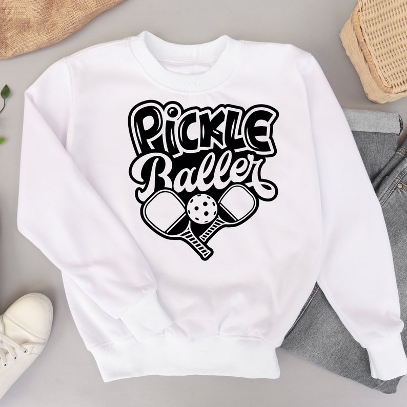 Pode incluir: Camisola branca com texto preto que diz "Pickle Baller" e um gr&aacute;fico de duas raquetes de pickleball e uma bola.