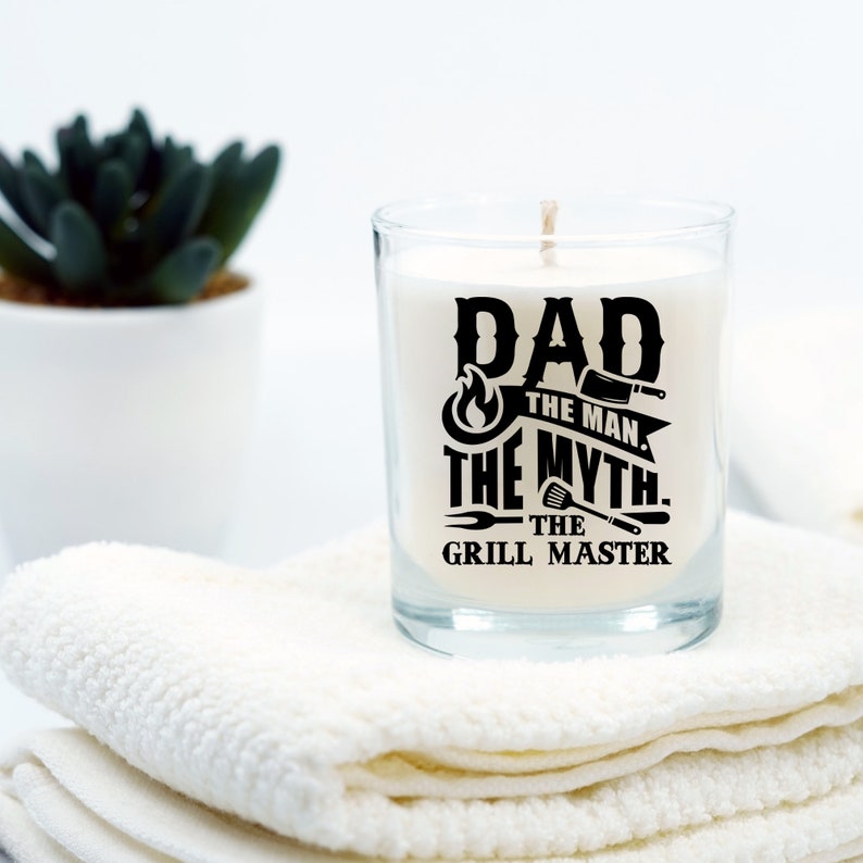 Puede incluir: Una vela blanca en un tarro de vidrio transparente con un dise&ntilde;o negro que dice "Dad The Man. The Myth. The Grill Master".