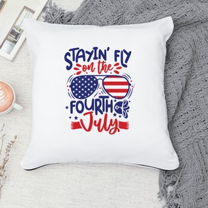 May include: White pillow with the text "Stayin' Fly on the Fourth of July" in blue and red lettering. The text is surrounded by stars and a pair of sunglasses with an American flag design.