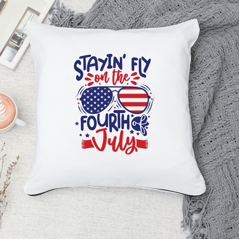 May include: White pillow with the text "Stayin' Fly on the Fourth of July" in blue and red lettering. The text is surrounded by stars and a pair of sunglasses with an American flag design.