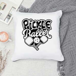 Pode incluir: Travesseiro branco com texto preto que diz "Pickle Baller" e um gr&aacute;fico de duas raquetes de pickleball e uma bola.