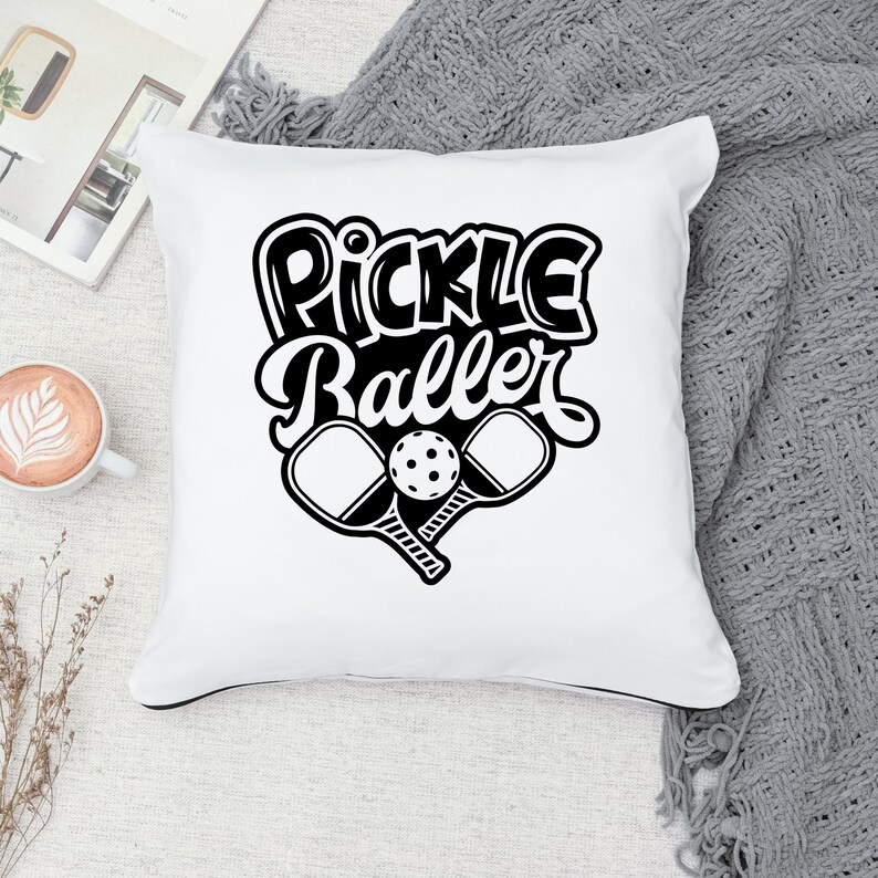 Pode incluir: Travesseiro branco com texto preto que diz "Pickle Baller" e um gr&aacute;fico de duas raquetes de pickleball e uma bola.