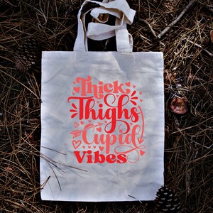 May include: White canvas tote bag with pink lettering that reads "Thick Thighs Cupid Vibes" with a pink arrow and hearts.