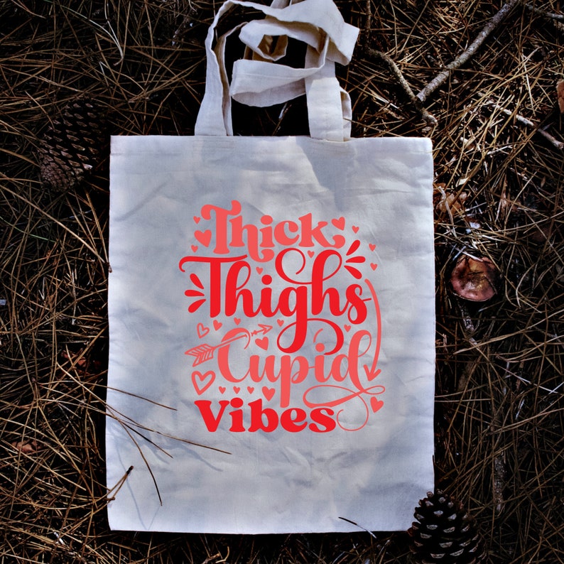 May include: White canvas tote bag with pink lettering that reads "Thick Thighs Cupid Vibes" with a pink arrow and hearts.