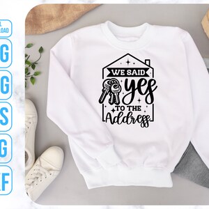Op de afbeelding: Wit sweatshirt met een zwart grafisch ontwerp van een huis met sleutels en de tekst "We said yes to the address".