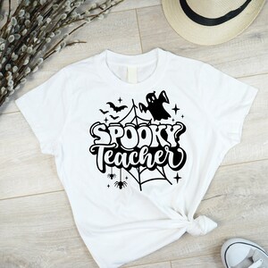 Pu&ograve; includere: Maglietta bianca con un design grafico nero di un fantasma, pipistrelli, una ragnatela e il testo "Spooky Teacher".