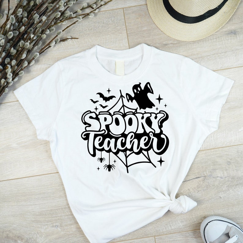 Pu&ograve; includere: Maglietta bianca con un design grafico nero di un fantasma, pipistrelli, una ragnatela e il testo "Spooky Teacher".