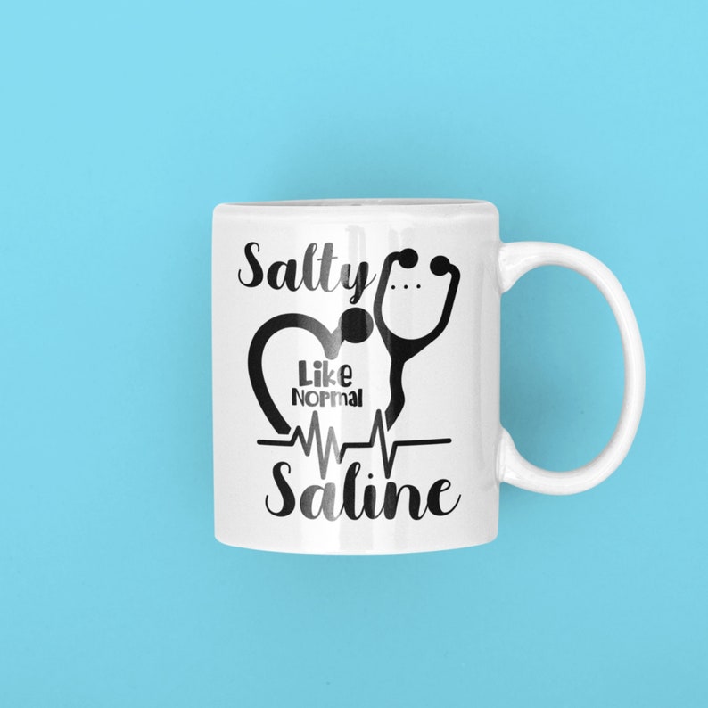 Peut inclure: Une tasse en c&eacute;ramique blanche avec un motif noir. Le motif repr&eacute;sente un st&eacute;thoscope, un c&oelig;ur et le texte "Salty Like Normal Saline".