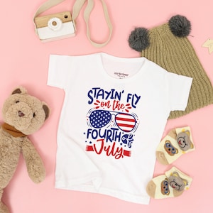 May include: White t-shirt with a blue and red graphic that says "Stayin' Fly on the Fourth of July" with sunglasses that have the American flag on them.