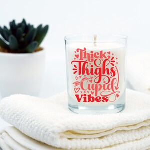 May include: A clear glass candle jar with a white candle inside. The candle has a red graphic that says "Thick Thighs Cupid Vibes" with hearts and an arrow.