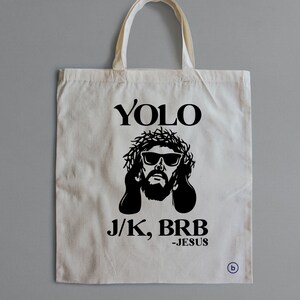 May include: A white tote bag with a black graphic of Jesus wearing sunglasses and a crown of thorns. The text "YOLO J/K, BRB -JESUS" is printed on the bag.