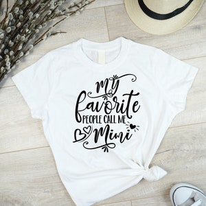 Puede incluir: Camiseta blanca con texto negro que dice "my favorite people call me Mimi" con dos corazones a cada lado de la palabra Mimi.