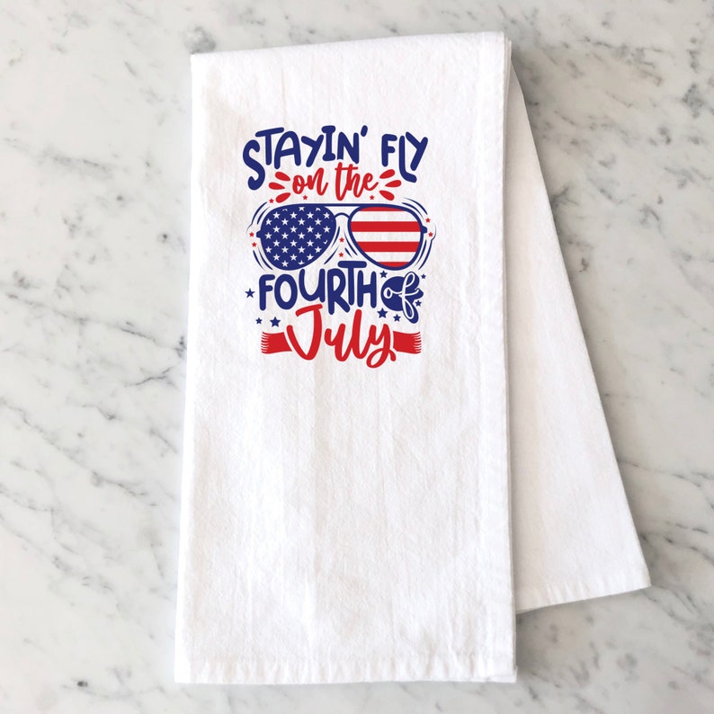May include: White kitchen towel with red, white, and blue text that reads "Stayin' Fly on the Fourth of July". The text is in a fun, playful font and features a pair of sunglasses with an American flag design.