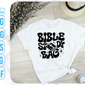 Op de afbeelding: Wit T-shirt met zwarte tekst die "Bible Study Bag" luidt in een retro lettertype. De tekst wordt omringd door sterren en een klein beeld van een gesloten boek met een kruis op de omslag.