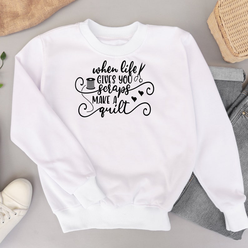 May include: White sweatshirt with black text that reads "When life gives you scraps make a quilt". The text is surrounded by decorative swirls and includes a sewing needle, scissors, and a spool of thread.